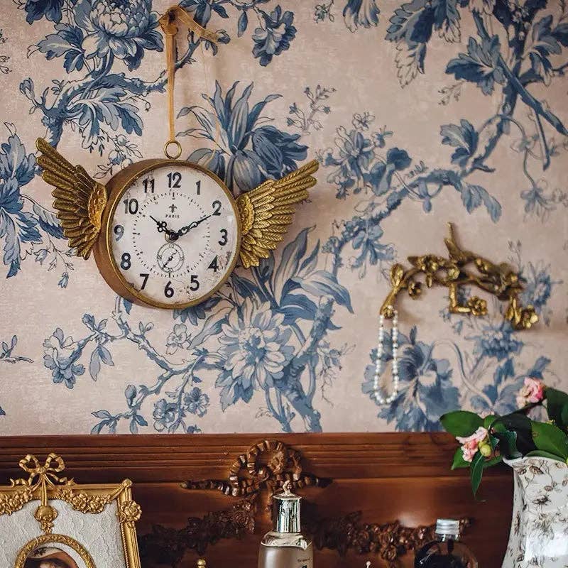 The IRON CLOCK IN GOLD FINISH by Vagabond Vintage hangs on floral wallpaper above a wooden dresser decorated with bottles, jewelry, and framed photos.