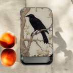 The Firefly Notes Black Crow Notions Tin – Handcrafted features a resin illustration of a black crow on a tree branch, displayed on a light surface beside two peaches, with sunlight casting shadows and highlighting the peaceful scene.