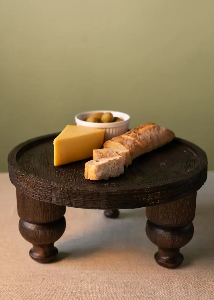 A 14in Wide Banjara Reclaimed Look Deep Brown/Black Rugged Wood Stand by Jamali Garden displays a wedge of yellow cheese, sliced baguette, and a white bowl of green olives against a neutral background.