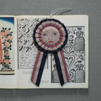 The Skippy Cotton Prize Ribbon is a handcrafted cotton-filled ornament featuring an embroidered face with pink cheeks and red lips, detailed black-and-white patterns, floral illustrations, and striped pink, black, and white ribbon tails.