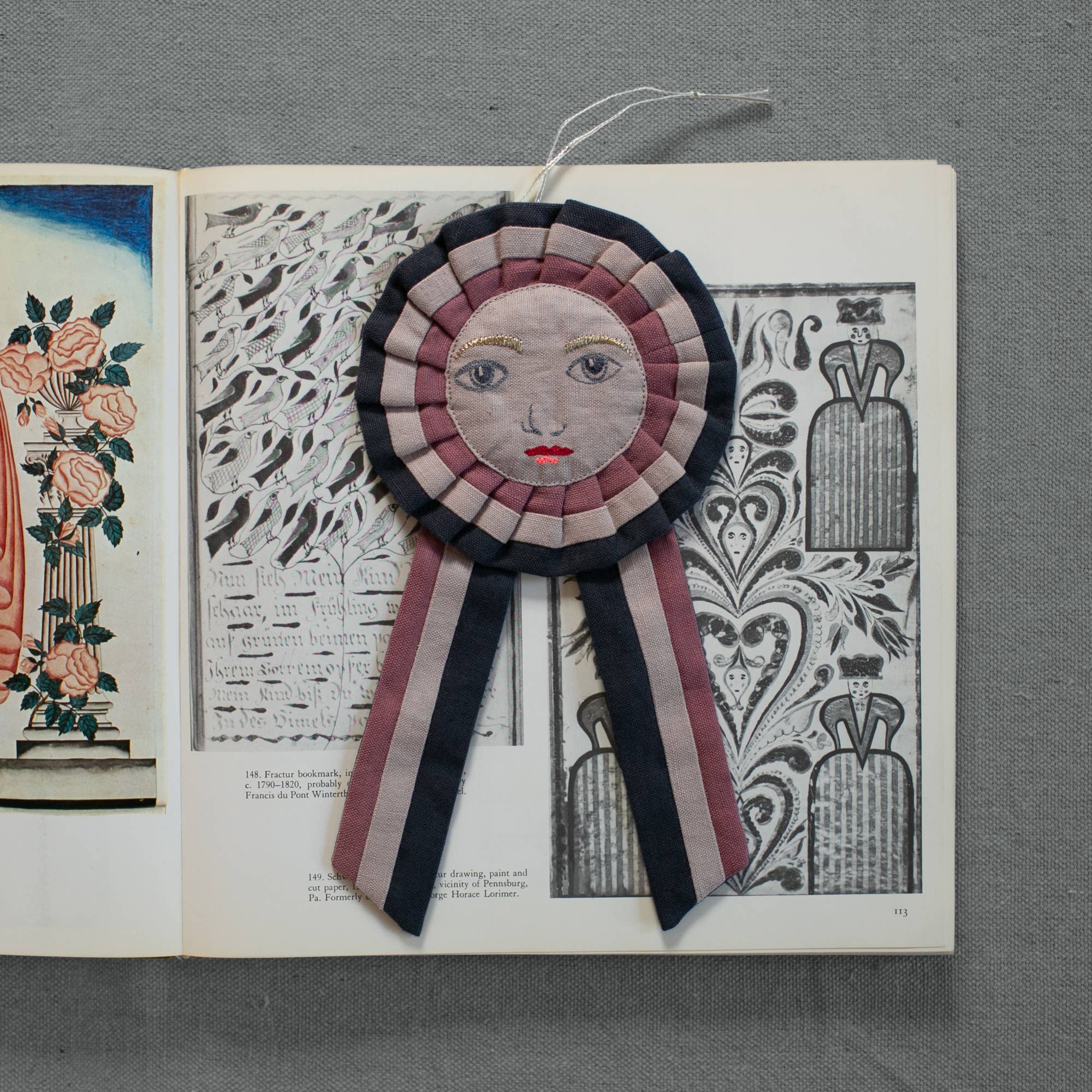 The Skippy Cotton Prize Ribbon is a handcrafted cotton-filled ornament featuring an embroidered face with pink cheeks and red lips, detailed black-and-white patterns, floral illustrations, and striped pink, black, and white ribbon tails.