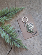 A green snail enamel keychain with a small leaf charm by ilikesara | art + goods is displayed on a brown "Animal Keychain" card atop wood, with green fern leaves nearby. Product: Metal enamel keychain / purse charm | snail.