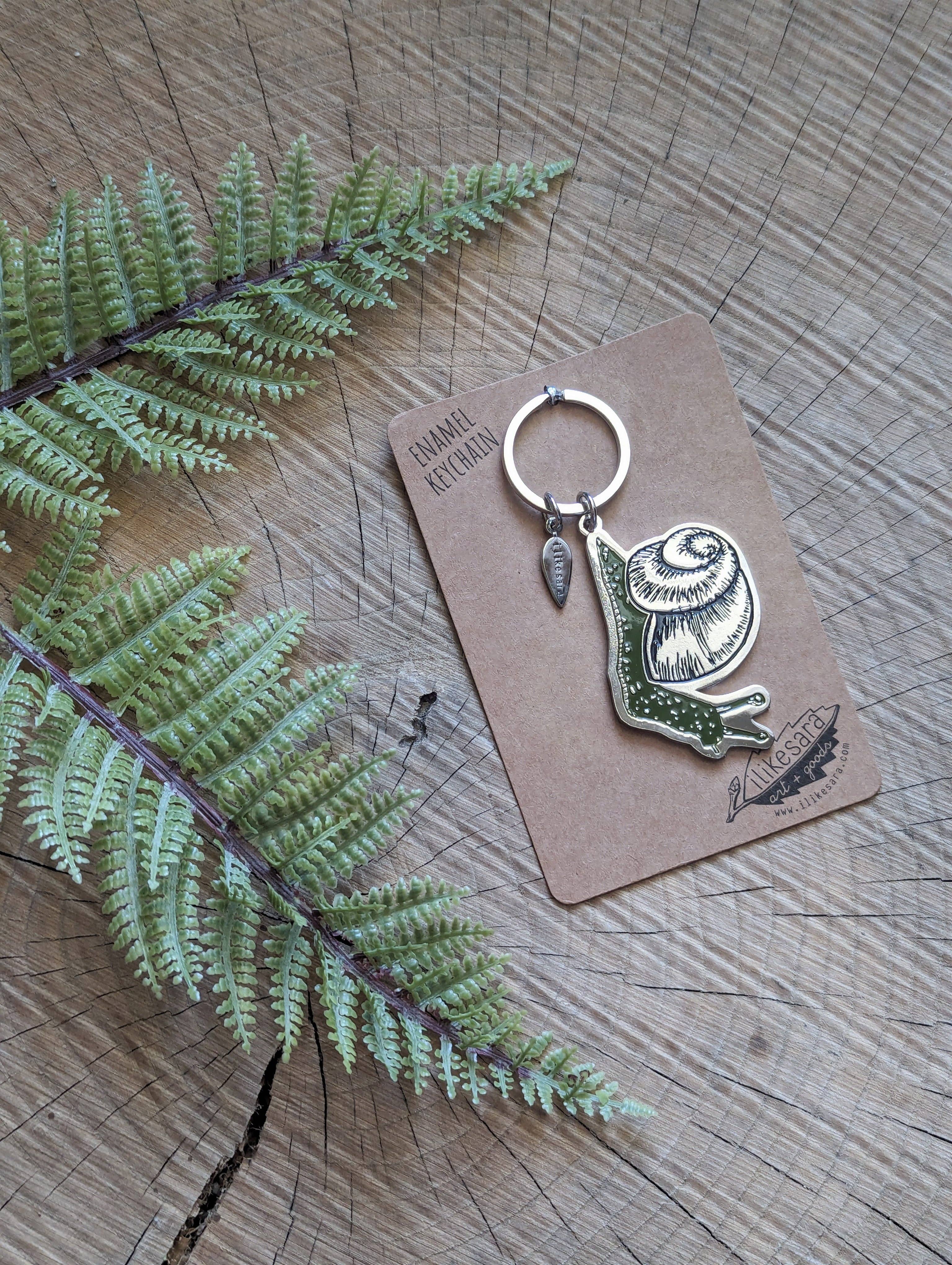 A green snail enamel keychain with a small leaf charm by ilikesara | art + goods is displayed on a brown "Animal Keychain" card atop wood, with green fern leaves nearby. Product: Metal enamel keychain / purse charm | snail.