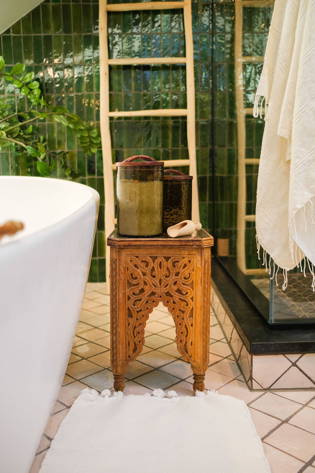 A carved wooden table displays Creative Women Handblown Hammered Glass Canisters: Smoke / Medium with soap beside a white tub, green tiles, wooden ladder, and white mat in a bright bathroom.