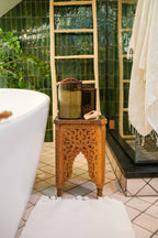 A carved wooden table displays Creative Women Handblown Hammered Glass Canisters: Smoke / Medium with soap beside a white tub, green tiles, wooden ladder, and white mat in a bright bathroom.