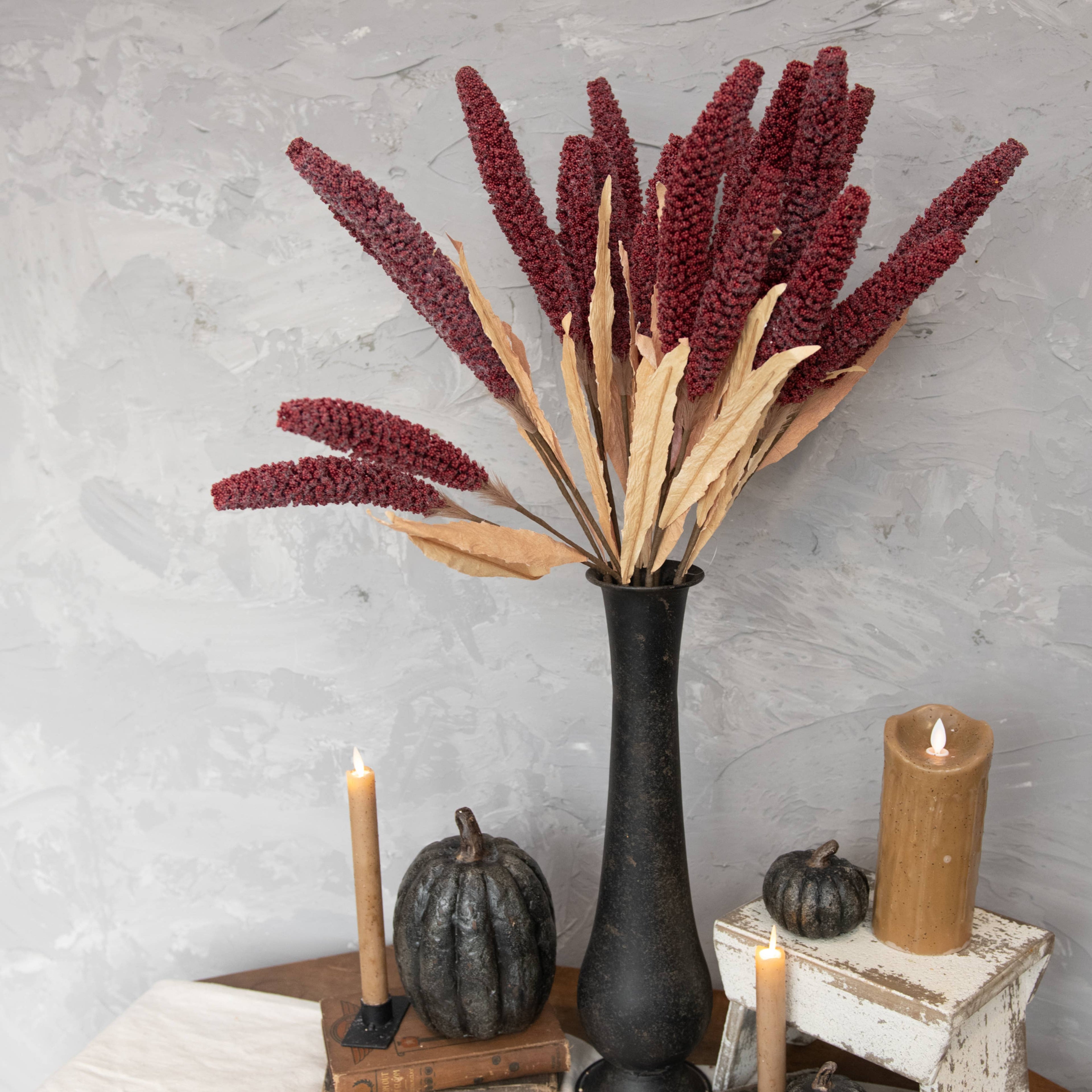 A 31" BURGUNDY SORGHUM SPRAY by Ragon House in a tall black vase is displayed on a table with candles, mini pumpkins, and vintage books, set against a textured gray wall.