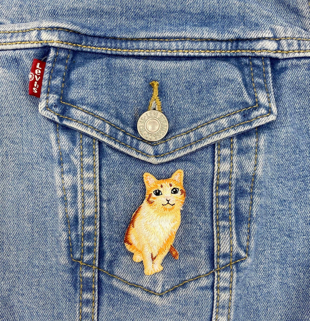 A Levis denim jacket pocket with a metal button features a Wholesale Applique Orange Tabby Cat embroidered iron on patch, showing a realistic kitten, sewn onto the pocket.