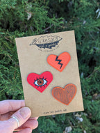 A hand holds a card labeled ilikesara | art + goods, showing the Iron-on patch | heart 3-pack - red: heart-shaped patches in orange with a black zigzag, red with an eye design, and orange with a wood grain pattern.