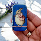 A hand holds the Firefly Notes Lady Bunbury, Bunny Notions Tin: Small, featuring a rabbit in a pink coat on blue. Slider-style bunny stitch markers and lavender sprigs are on a white wooden surface in the background.