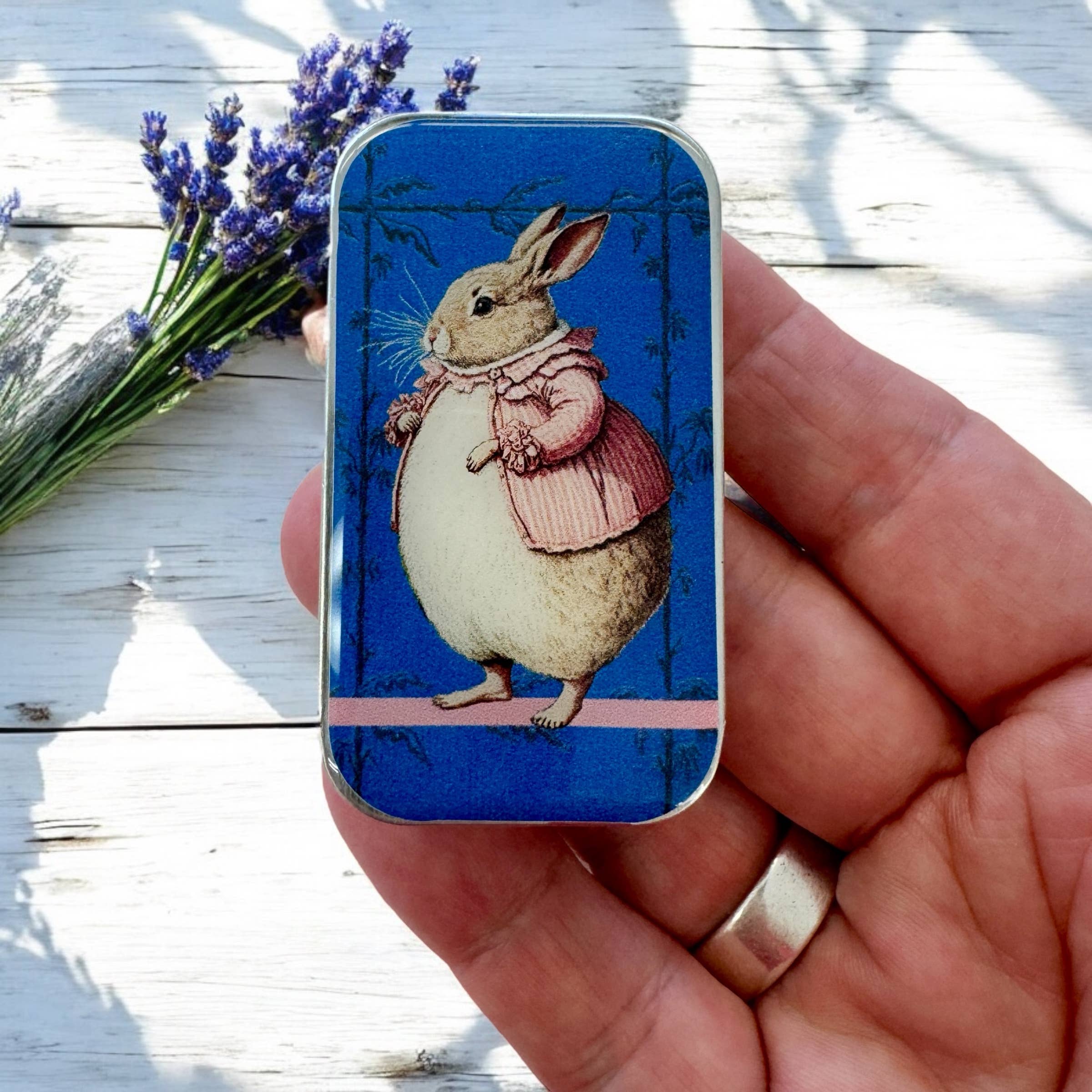A hand holds the Firefly Notes Lady Bunbury, Bunny Notions Tin: Small, featuring a rabbit in a pink coat on blue. Slider-style bunny stitch markers and lavender sprigs are on a white wooden surface in the background.