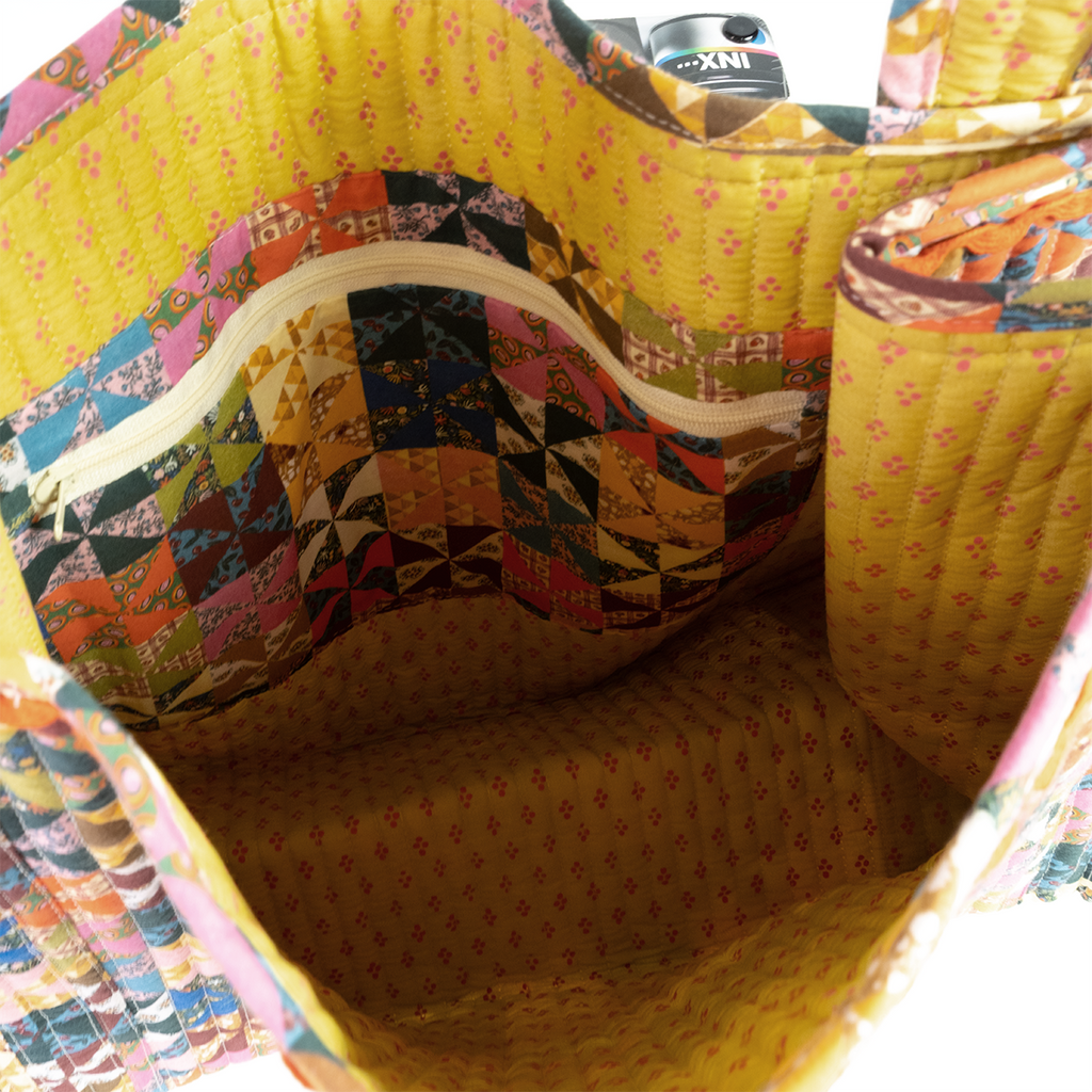 A close-up of the Calico Quilted Tote by Handker Bandanas reveals its colorful patchwork, yellow interior, inner zippered pocket, and a camera peeking out.