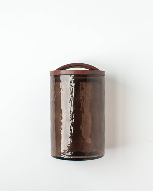 The Handblown Hammered Glass Canisters: Smoke / Medium by Creative Women is a tall, cylindrical recycled glass canister with a hand-carved wooden lid and handle—ideal for airtight kitchen storage.