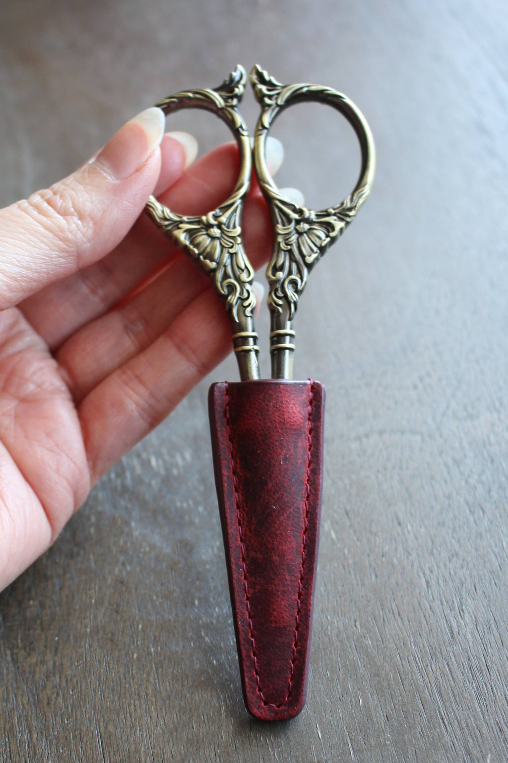 A hand holds ornate embroidery scissors, partially covered with the NNK Press Leather Scissor Sheath, set against a wooden background—an ideal accessory to protect your embroidery tools.