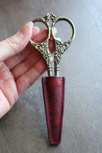 A hand holds ornate embroidery scissors, partially covered with the NNK Press Leather Scissor Sheath, set against a wooden background—an ideal accessory to protect your embroidery tools.