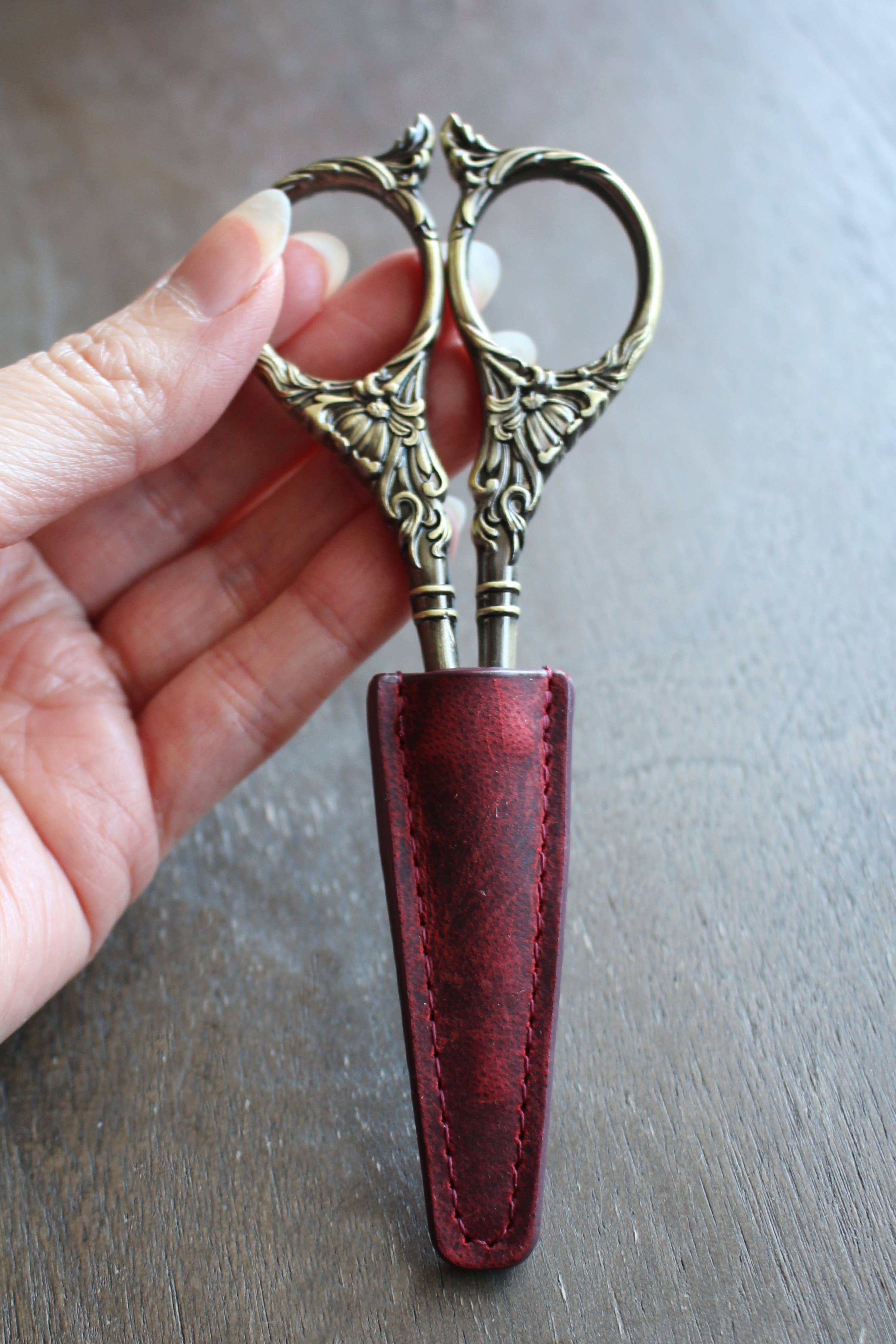 A hand holds ornate embroidery scissors, partially covered with the NNK Press Leather Scissor Sheath, set against a wooden background—an ideal accessory to protect your embroidery tools.