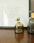 A delicate bloom rests in the Bella Handblown Hammered Glass Bud Vase: Blush by Creative Women, placed on a wooden surface beside a larger jar and a framed handwritten letter in the background.