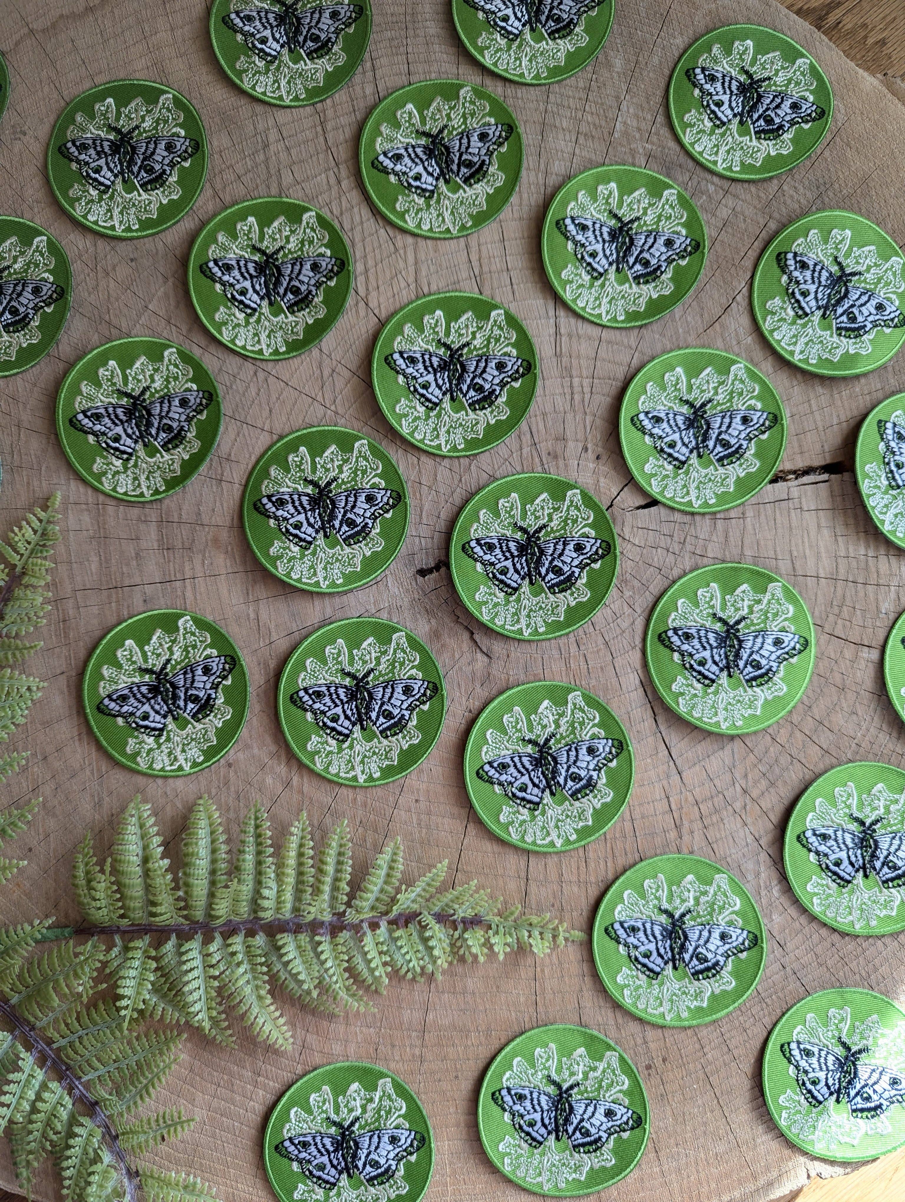 The Iron-on patch | polyphemus moth with lichen - green by ilikesara | art + goods features a detailed moth and lichen design, displayed on a tree-ringed surface with fern leaves accenting the nature-inspired aesthetic.