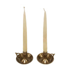 Two tall, cream-colored tapers are set in matching Brighton Bun Traveling Chambersticks by Madison Bay Co.—an elegant antique revival with small handles, displayed on a plain white background.