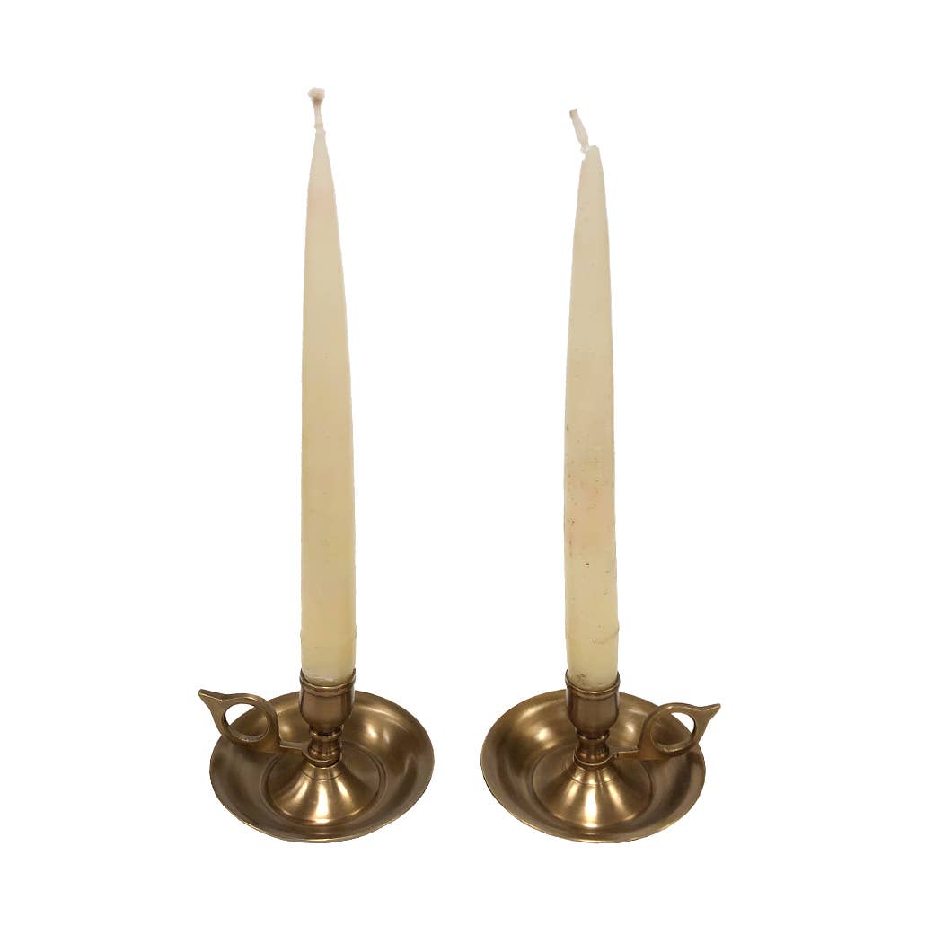 Two tall, cream-colored tapers are set in matching Brighton Bun Traveling Chambersticks by Madison Bay Co.—an elegant antique revival with small handles, displayed on a plain white background.