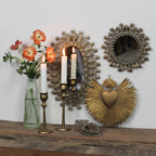 A wooden shelf displays two lit white candles in HomArt Sonrisa Brass Taper Holders, a clear vase with orange flowers, and ornate gold and silver mirrors and heart wall decor hanging above.