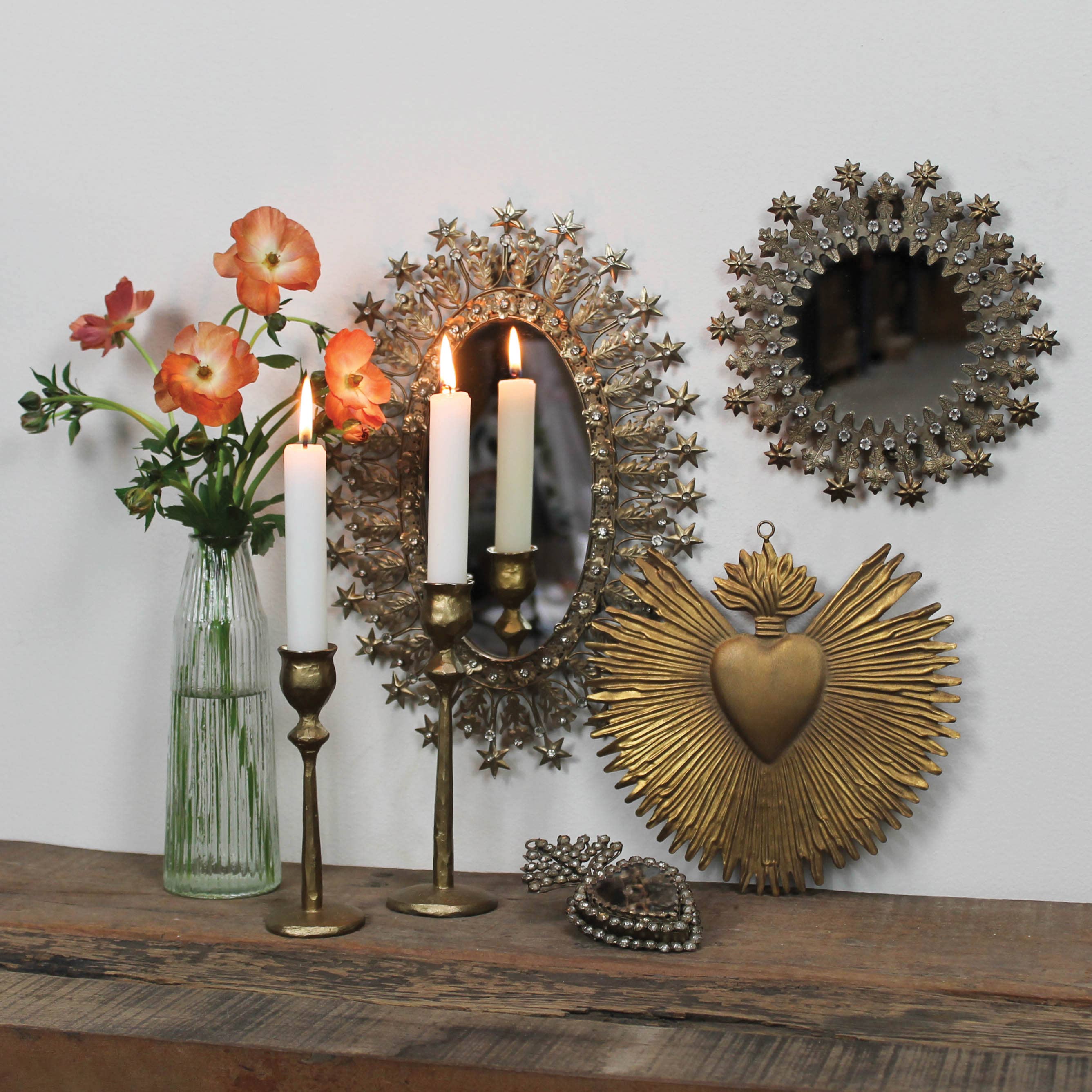 A wooden shelf displays two lit white candles in HomArt Sonrisa Brass Taper Holders, a clear vase with orange flowers, and ornate gold and silver mirrors and heart wall decor hanging above.