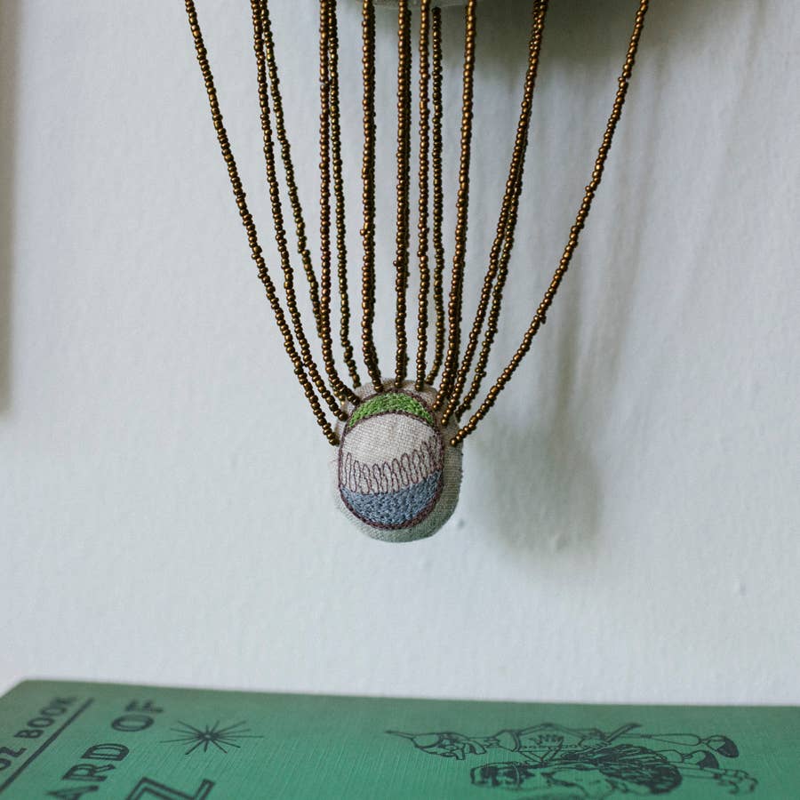 The Skippy Cotton "Wonderful Wizard / Balloon, Oz Wallhanging" features a fabric ball suspended by beaded strings above a green book, creating whimsical embroidered wall art or an ornament.