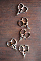 Five NNK Press Mini Embroidery Scissors in Antique Gold, featuring stainless steel blades and intricate vintage-style handles, are arranged in a zigzag pattern on a wooden surface.