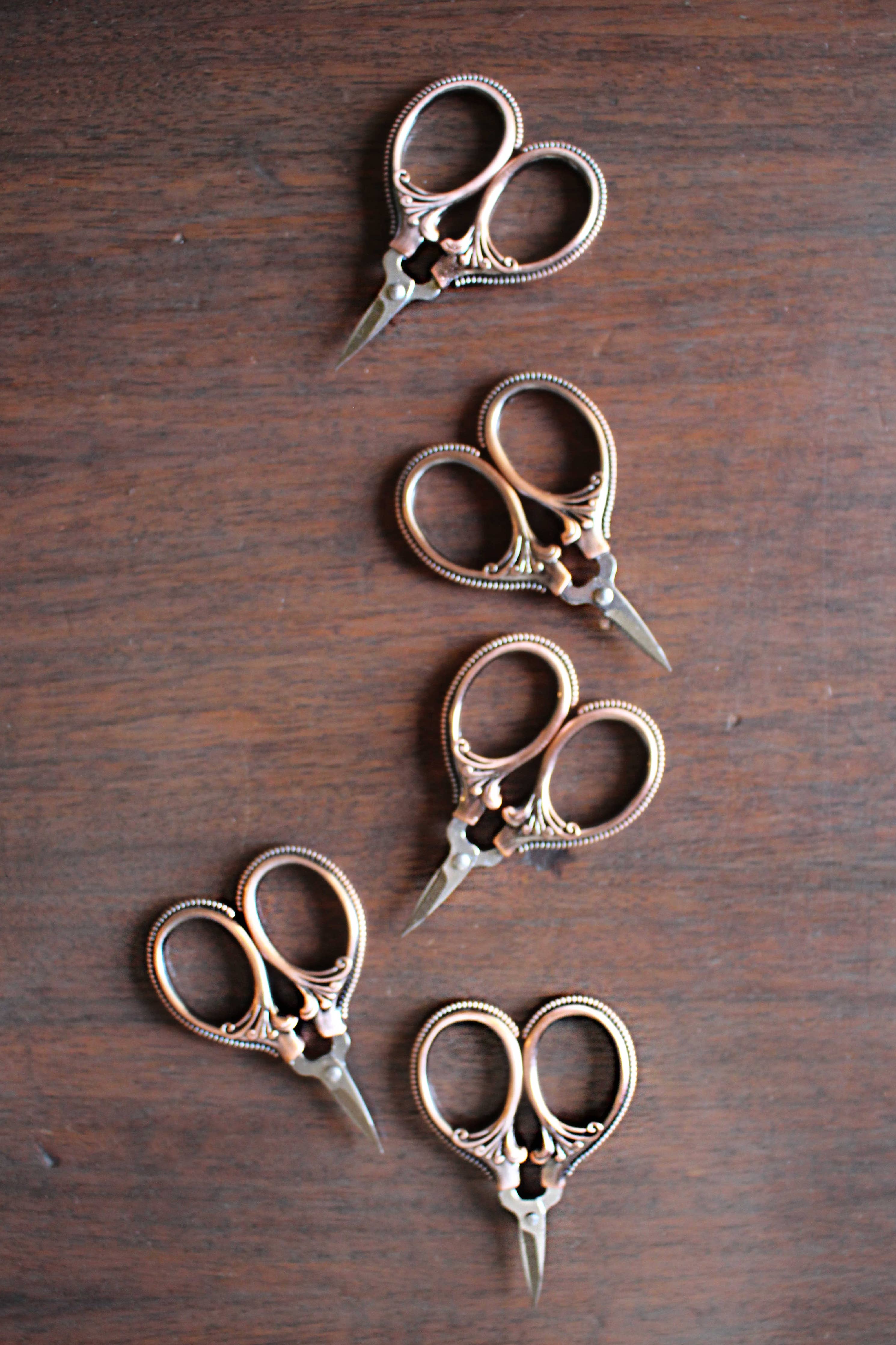 Five NNK Press Mini Embroidery Scissors in Antique Gold, featuring stainless steel blades and intricate vintage-style handles, are arranged in a zigzag pattern on a wooden surface.