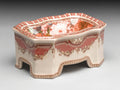 The AA Importing Fruit and Flower Porcelain Compote features ornate red and white floral patterns, curved edges, and four short legs, creating a decorative accent against a neutral background.