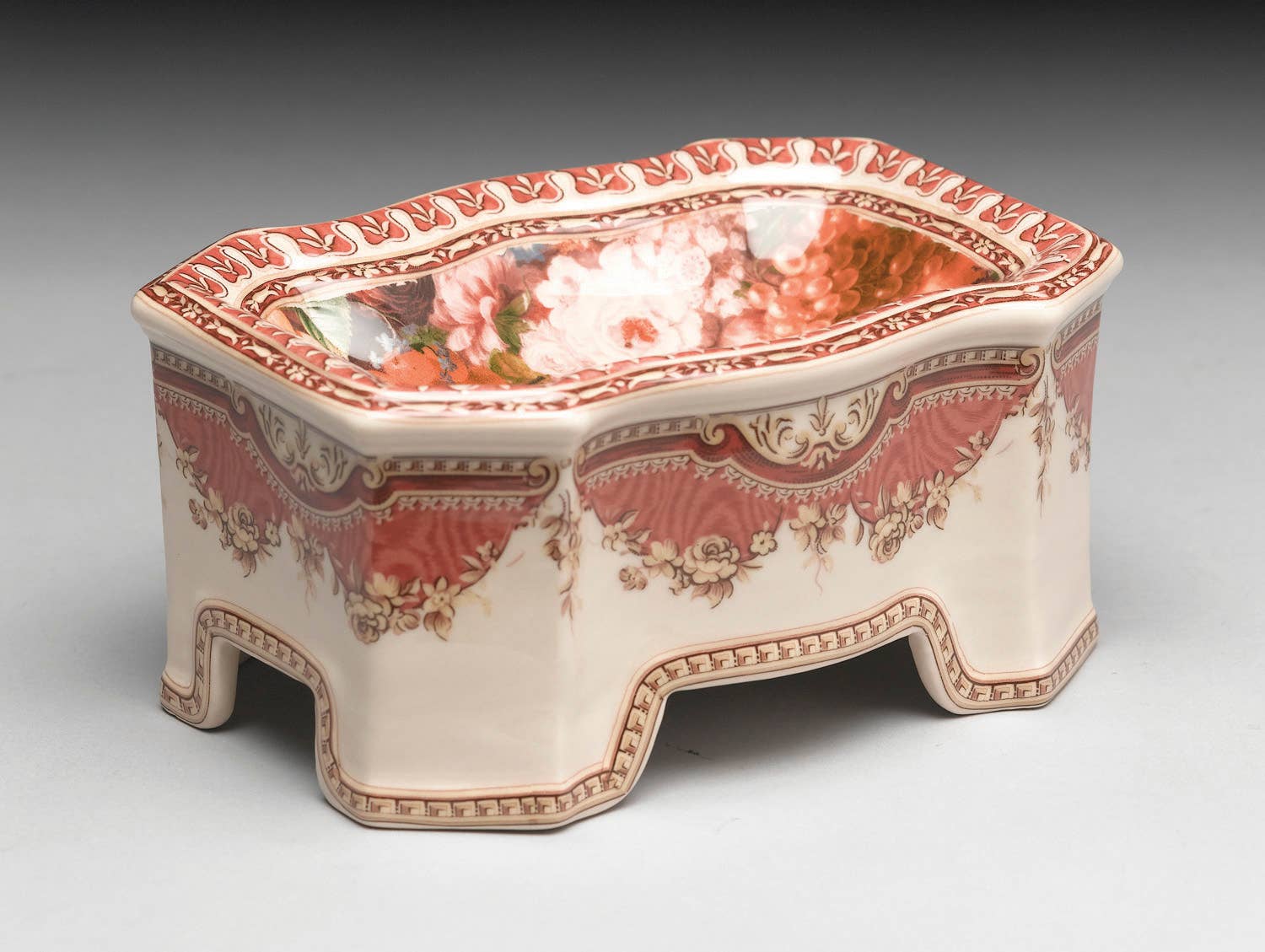 The AA Importing Fruit and Flower Porcelain Compote features ornate red and white floral patterns, curved edges, and four short legs, creating a decorative accent against a neutral background.