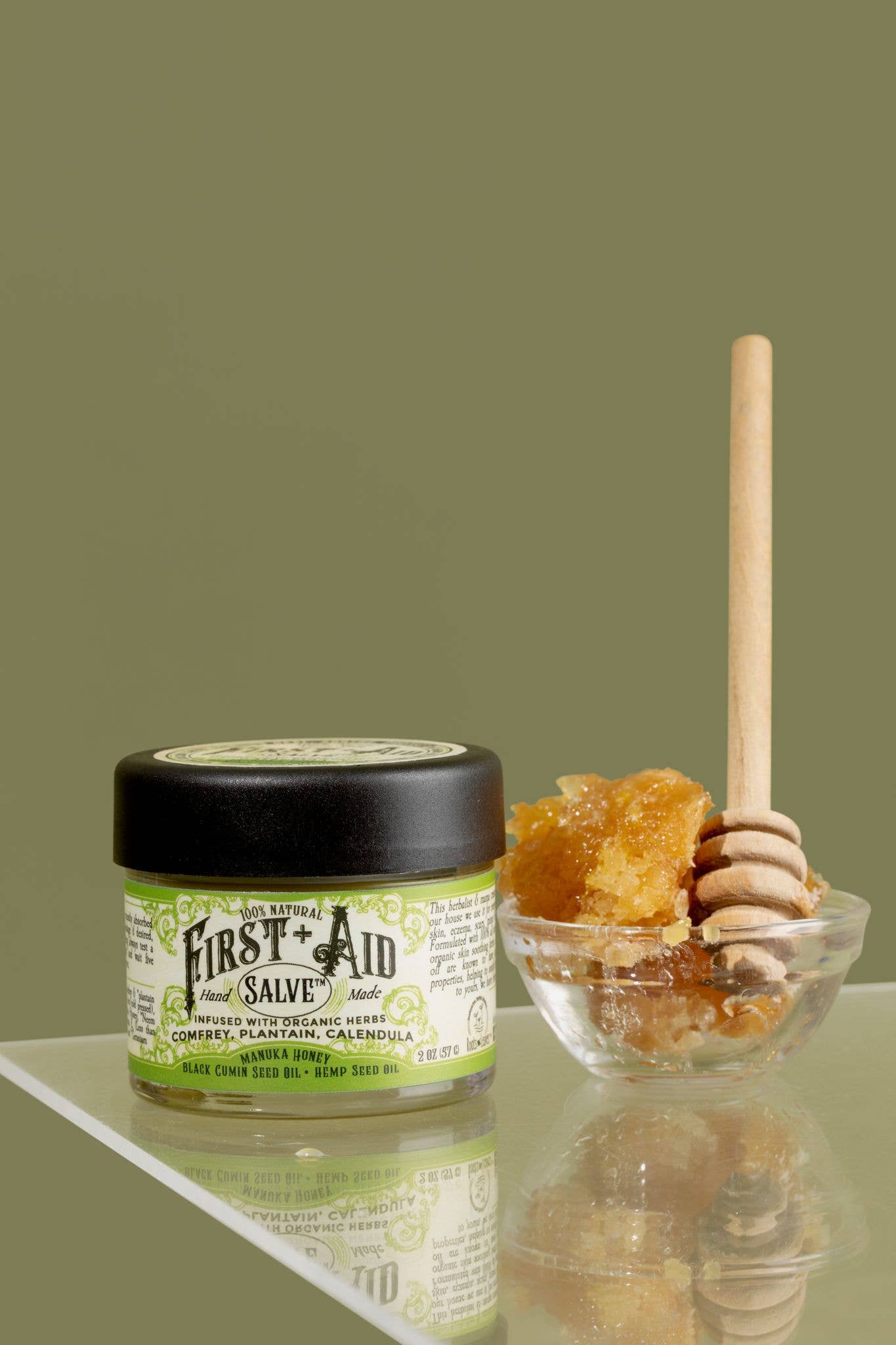 A jar of Roots And Leaves First Aid Salve sits beside a small glass bowl of honeycomb with a wooden dipper, all arranged on a reflective surface against an olive green background.