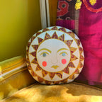 The Skippy Cotton Bright Neon Sun Pillow, a handmade cotton-filled cushion with yellow and red rays and rosy cheeks, sits on a mustard chair beside vibrant fabric—perfect for adding a whimsical touch to your space.