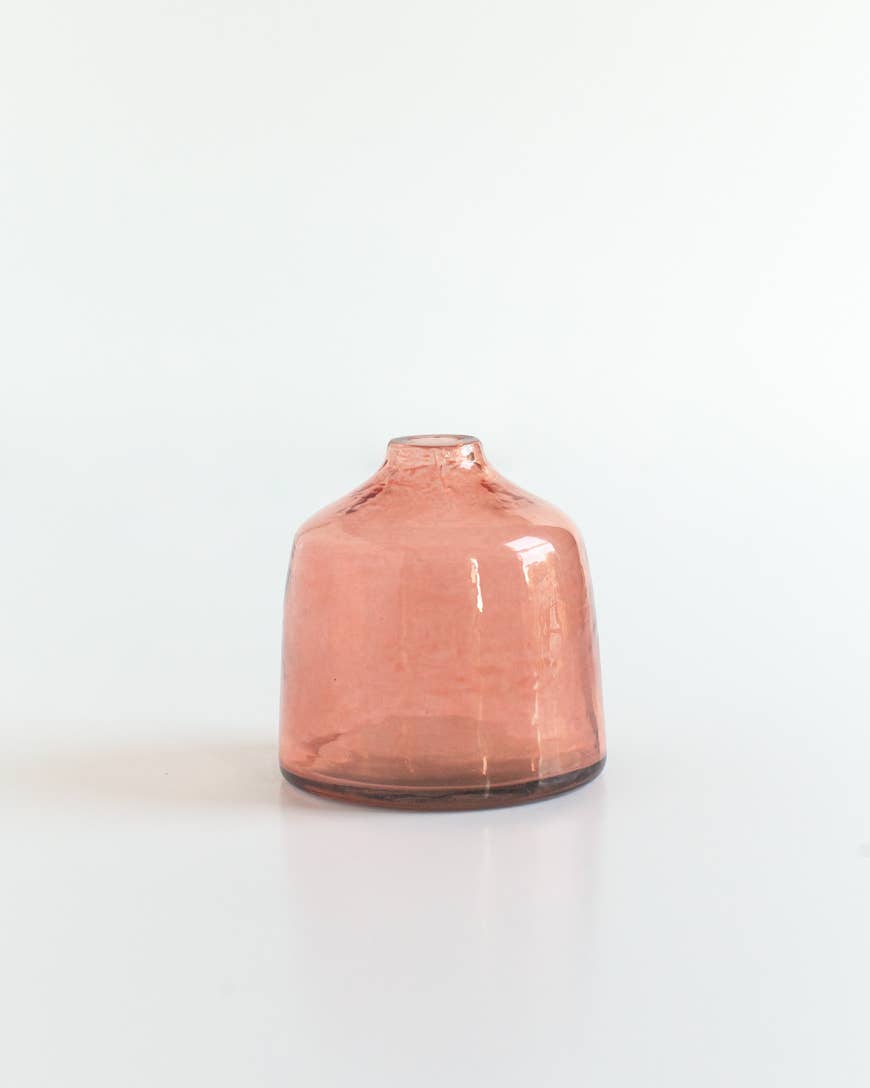 The Bella Handblown Hammered Glass Bud Vase: Blush by Creative Women, a small round pink recycled glass vase with a narrow opening, sits on a white surface against a plain white background.