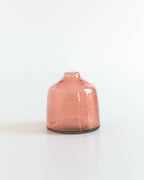 The Bella Handblown Hammered Glass Bud Vase: Blush by Creative Women, a small round pink recycled glass vase with a narrow opening, sits on a white surface against a plain white background.