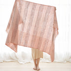 A barefoot person stands on a light-toned floor, holding up the Variously Silica Rose Throw in Shades of Salmon, featuring gray stripes and a salmon rose hue; the large, handwoven textile obscures their upper body and face, with sheer curtains in the background.
