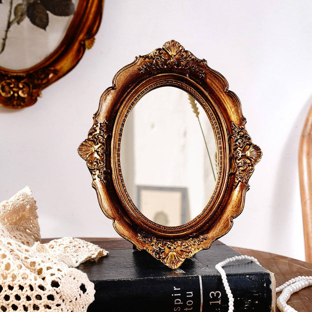 The Vagabond Vintage Vintage Style Ornate Oval Table Mirror: Medium, with a decorative gold frame, sits on a wooden surface beside a crocheted cloth and black book; a blurred framed picture hangs on the white wall behind.