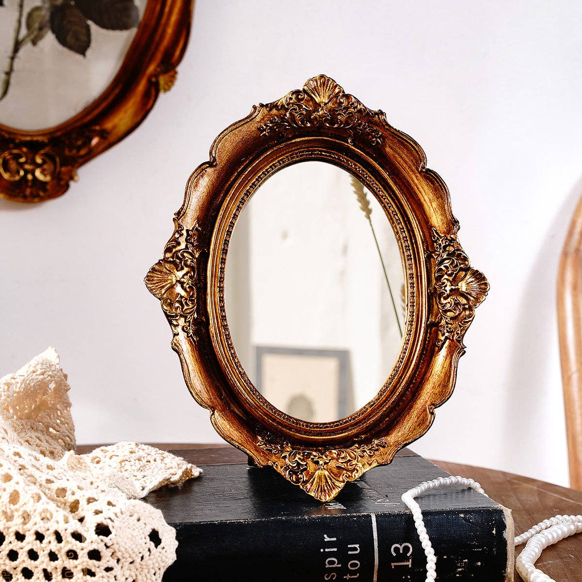 The Vagabond Vintage Vintage Style Ornate Oval Table Mirror: Medium, with a decorative gold frame, sits on a wooden surface beside a crocheted cloth and black book; a blurred framed picture hangs on the white wall behind.