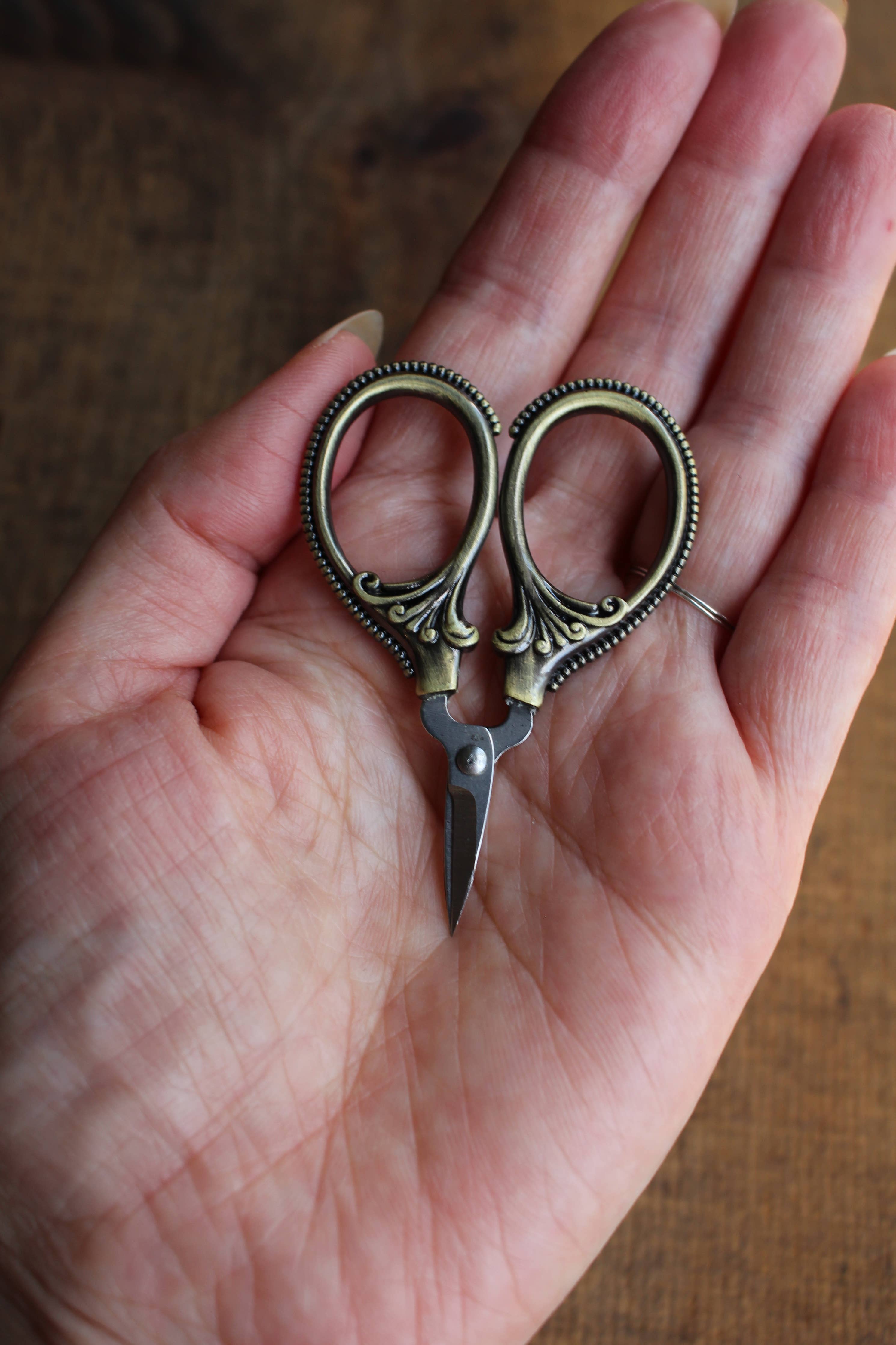 A hand holds NNK Press Mini Embroidery Scissors: Antique Gold, featuring ornate handles and stainless steel blades—perfect compact sewing snips for travel notions—set on a wooden background.