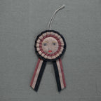 The Skippy Cotton Prize Ribbon - Cotton Filled Ornament features dark, light pink, and beige stripes with an embroidered face and two tails, creating a charming handcrafted accent against textured gray fabric.