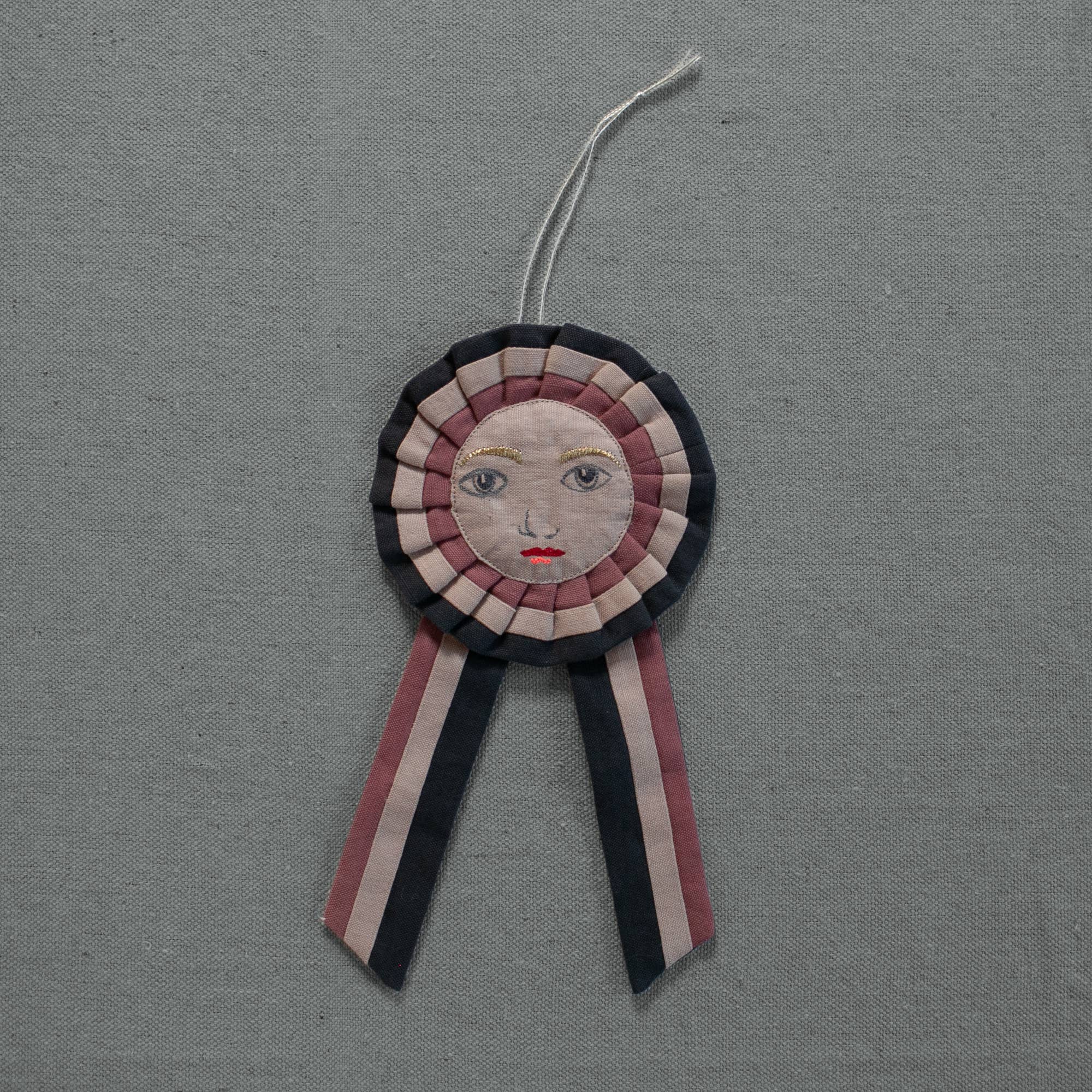 The Skippy Cotton Prize Ribbon - Cotton Filled Ornament features dark, light pink, and beige stripes with an embroidered face and two tails, creating a charming handcrafted accent against textured gray fabric.