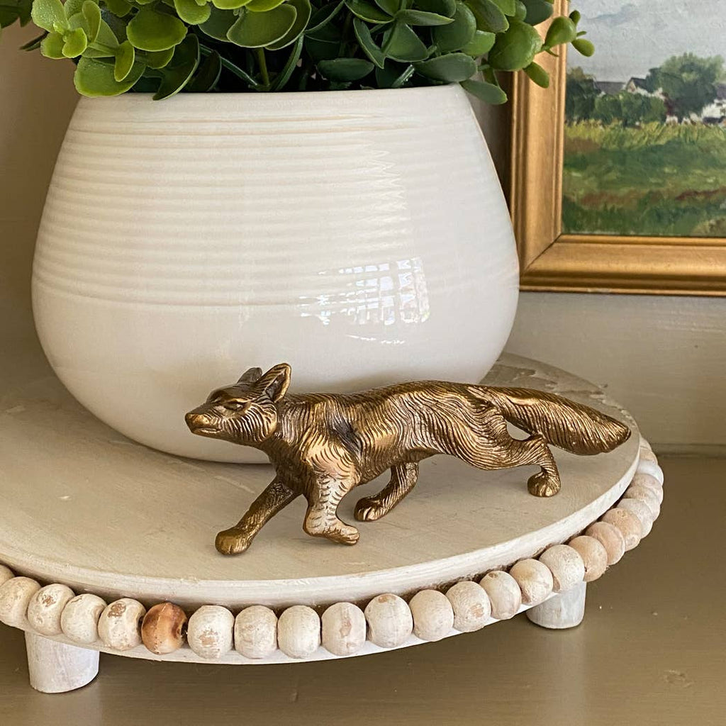 The Solid Antiqued Brass Running Fox Tabletop Desk Decor by Madison Bay Co. sits on a round, beaded white tray with a potted plant and a framed landscape painting in the background.
