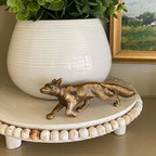 The Solid Antiqued Brass Running Fox Tabletop Desk Decor by Madison Bay Co. sits on a round, beaded white tray with a potted plant and a framed landscape painting in the background.