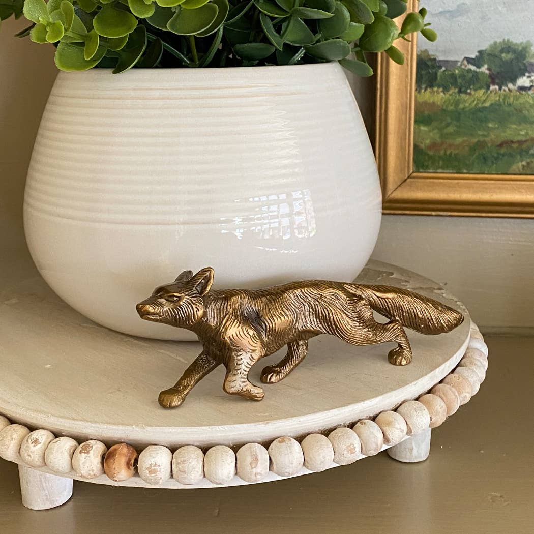 The Solid Antiqued Brass Running Fox Tabletop Desk Decor by Madison Bay Co. sits on a round, beaded white tray with a potted plant and a framed landscape painting in the background.