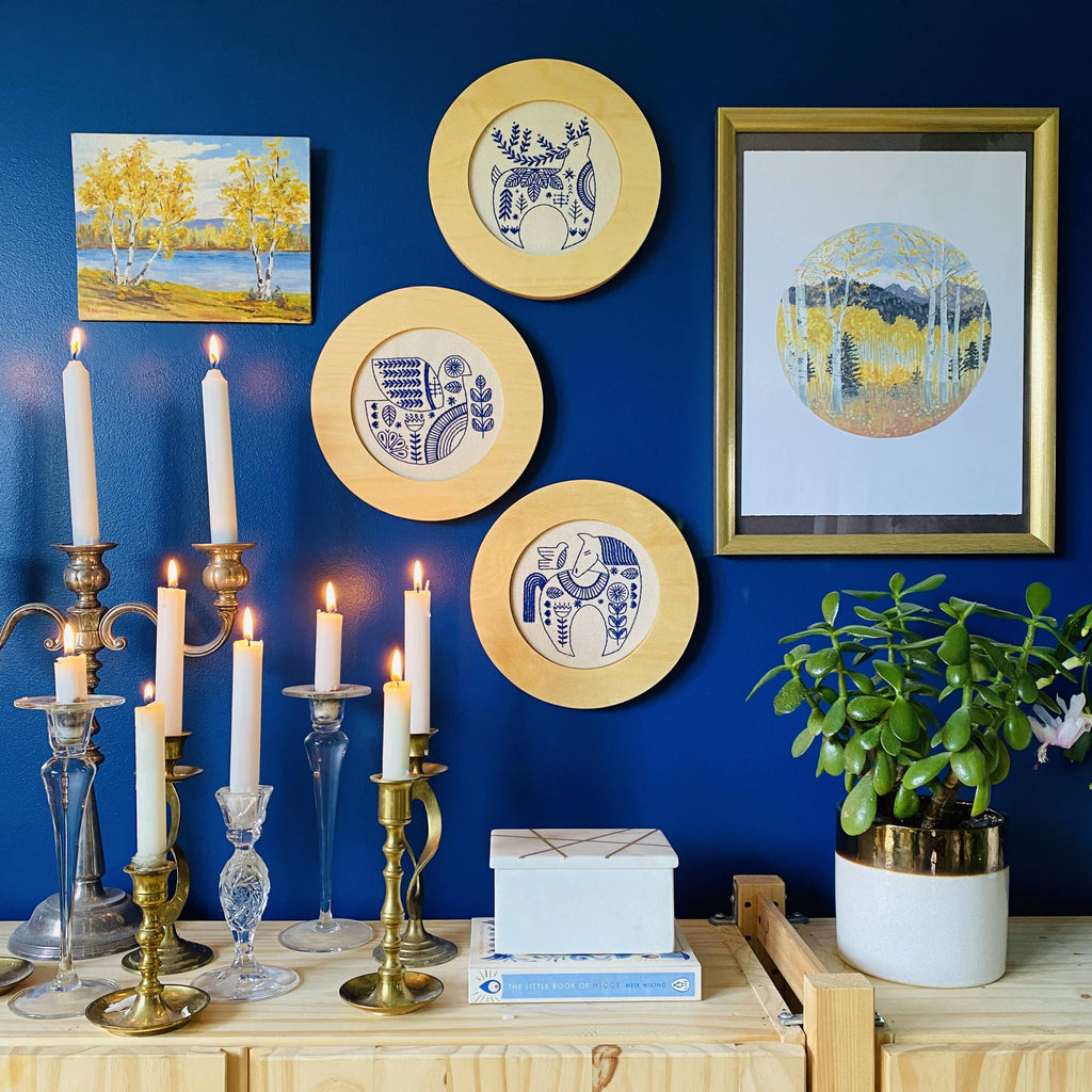 A blue wall features three decorative plates, a framed picture, and a painting. On the wooden shelf below are potted plant, stacked boxes, the Hygge Dove Complete Embroidery Kit by Hook, Line & Tinker, and lit candles in glass and brass holders.