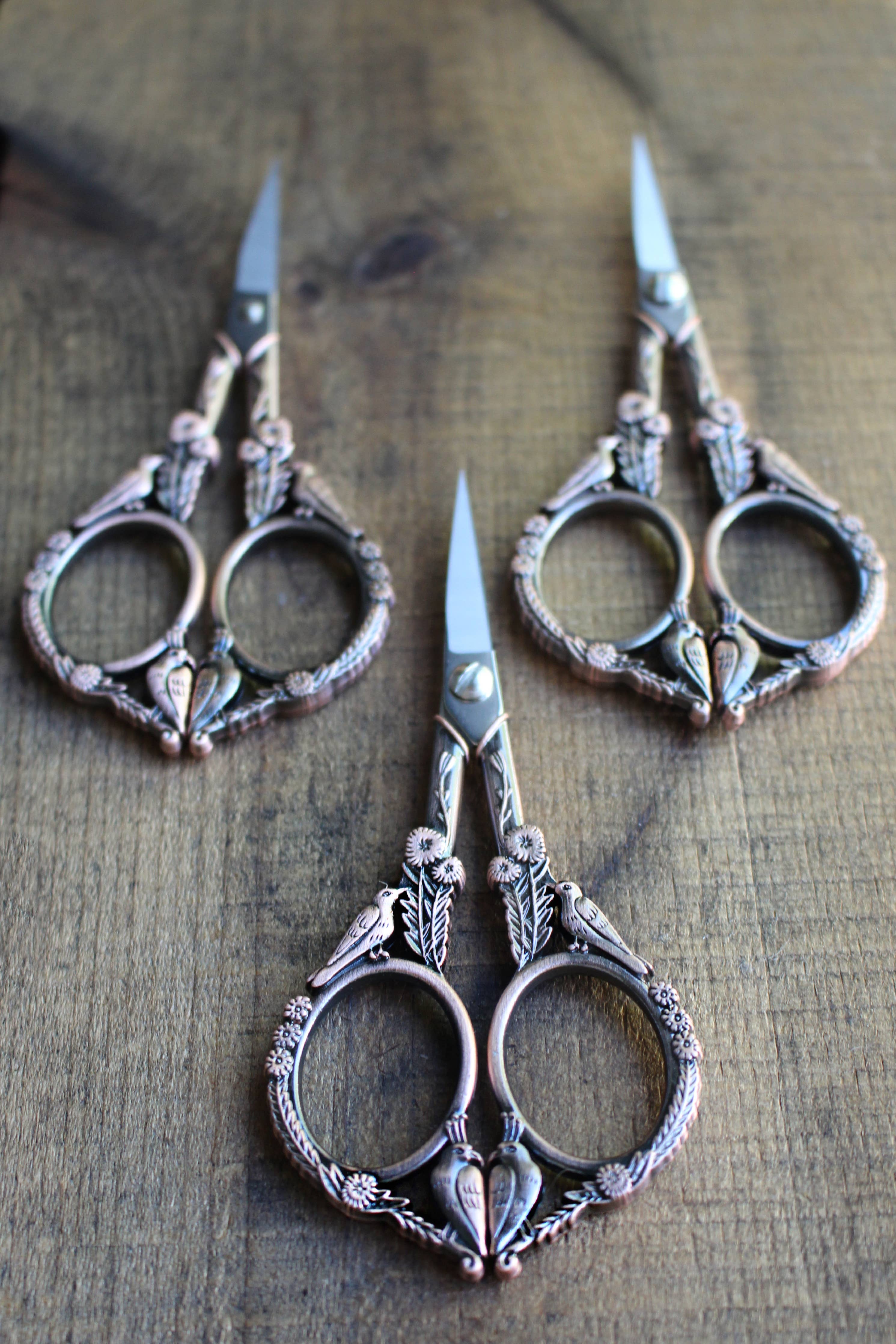 Three Feathered Friends Scissors: Antique Gold by NNK Press, with ornate handles and stainless steel blades, are arranged on a wooden surface, their tips fanned out in an elegant display.