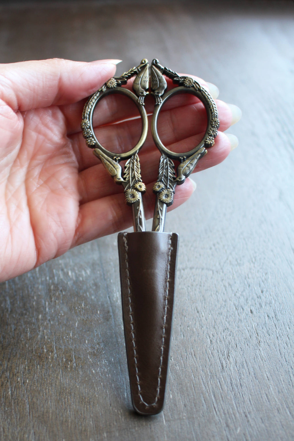 A hand displays ornate embroidery scissors with leaf and bird motifs, partially sheathed in the NNK Press Leather Scissor Sheath, set against a wooden surface.