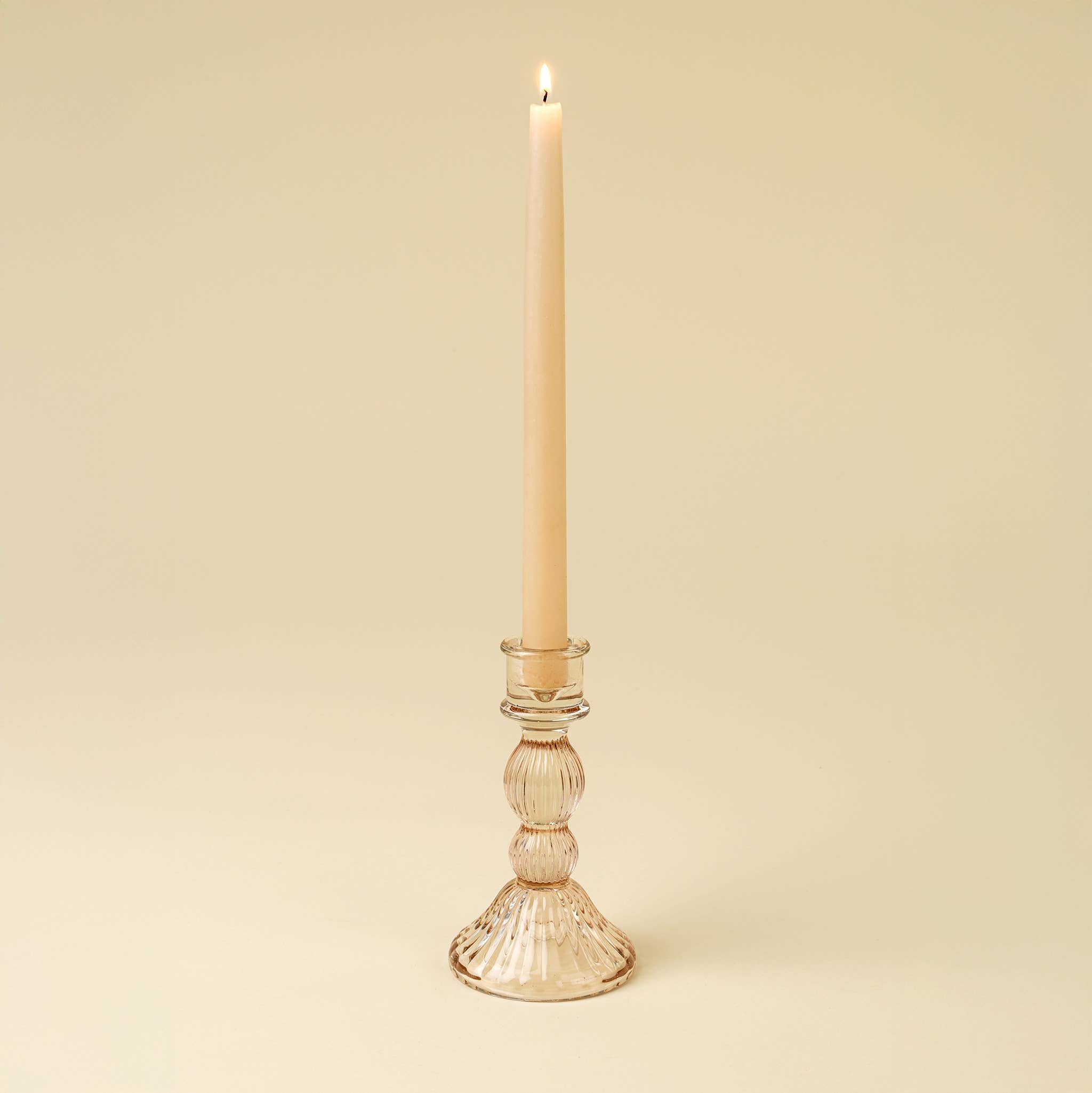 A lit beige taper candle stands in the Indaba Trading Callista Glass Candleholder Large, Blush, against a plain light beige background, bringing vintage-inspired style to your decor.