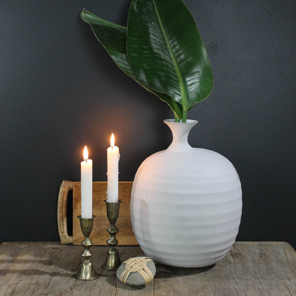 A white ceramic vase with large green leaves sits on a wooden surface beside two lit candles in HomArt's Haiku Taper Holder, Brass - Med, and a small decorative piece, all set against a dark background.