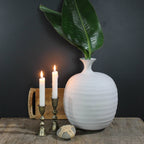 A white ceramic vase with large green leaves sits on a wooden surface beside two lit candles in HomArt's Haiku Taper Holder, Brass - Med, and a small decorative piece, all set against a dark background.