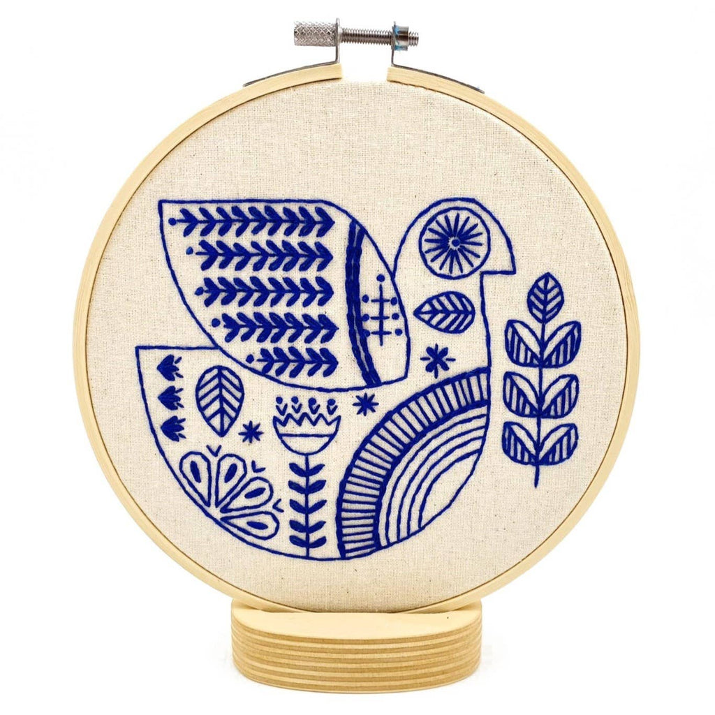The Hygge Dove Complete Embroidery Kit by Hook, Line & Tinker features a blue-stitched stylized bird with geometric patterns and leaves on beige fabric. Includes an upright embroidery hoop on a wooden base, ideal for display.